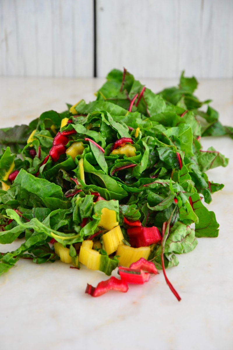A bunch of chopped Swiss card on a marble countertop with veggies mixed in.