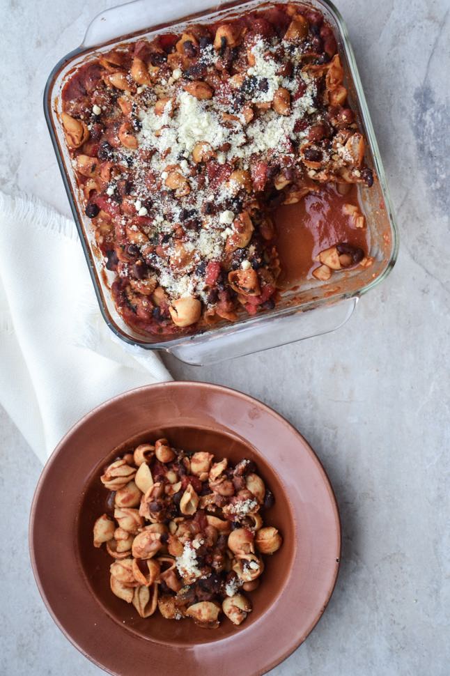 Looking down on a glass casserole dish filled with pasta shells, beans, and red chili, and a serving plated beside it. 