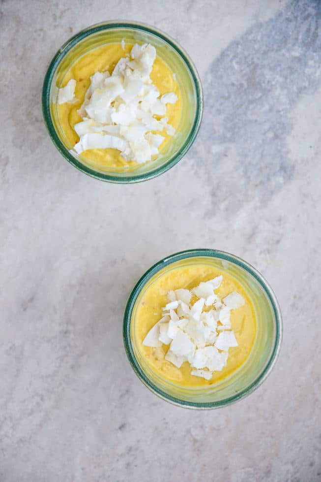 Easy 5 ingredient Mango Pudding recipe that's topped with coconut flakes in beautiful glasses on a marble table.