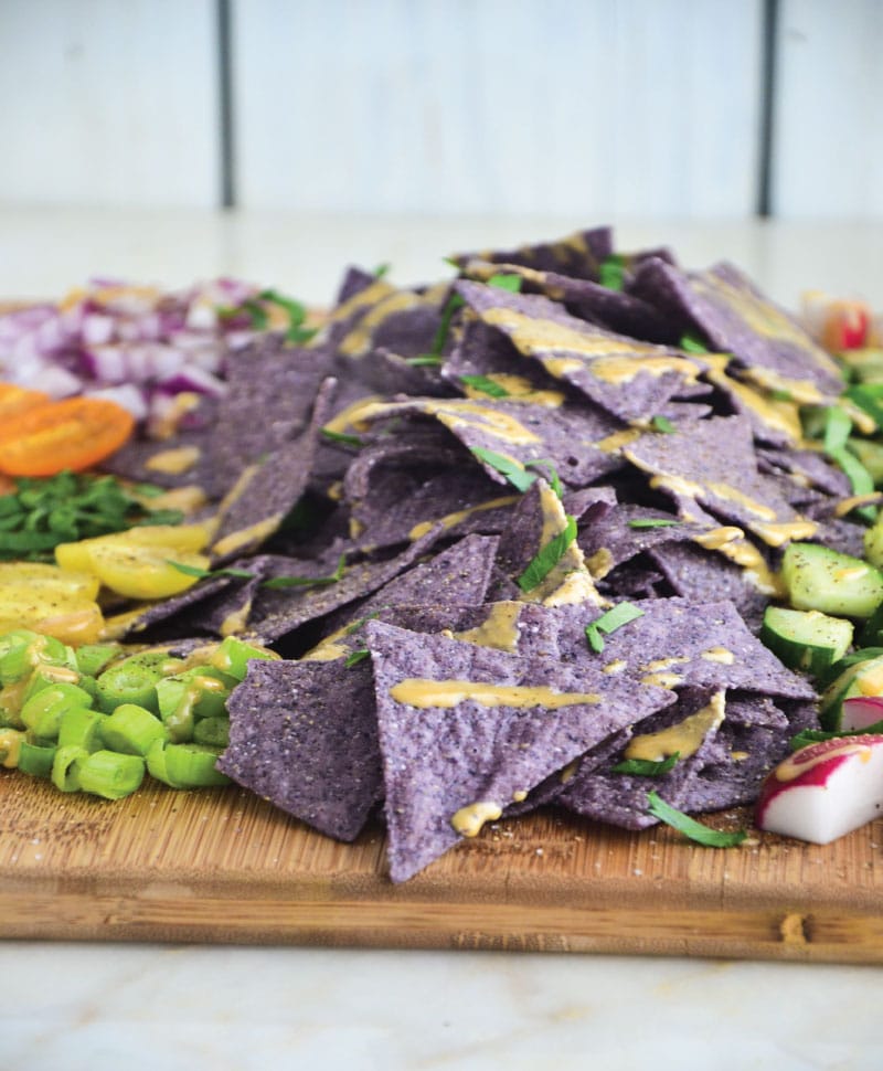 Blue gluten-free chips with tahini sauce and toppings on a wooden cutting board.