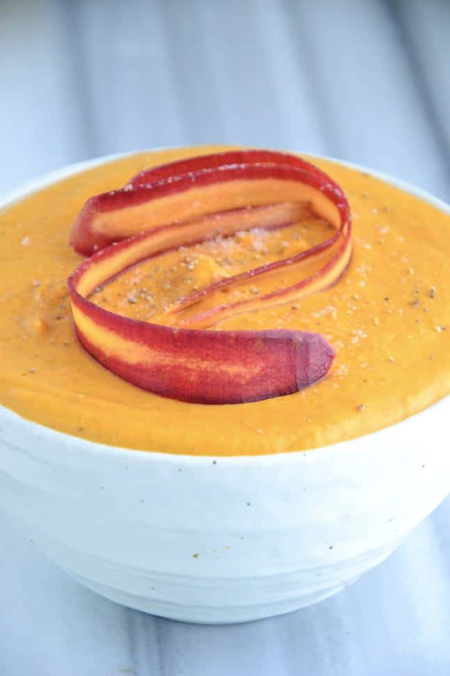 A white bowl brimming with beautiful, creamy orange soup, topped with a rainbow carrot ribbons.